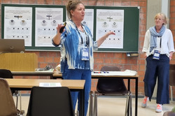Frauke Mörik (Frau im mittleren Alter, blau gekleidet) steht vor drei Postern in einem Seminarraum. Hinten ist eine blonde Frau und vier Poster auf einer Tafel zu erkennen.