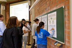 In einem Seminarraum stehen mehrere Personen rechts vor einer Tafel, auf der Poster angebracht sind. Sie schreiben etwas darauf, sehen es sich an oder betrachten es. Hinten ist eine projizierte Folie zu erkennen.