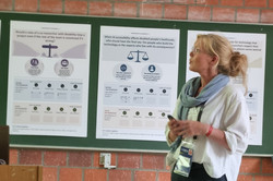 Susane Dirks (die blonde Frau im mittleren Alter) steht vor drei Postern und schaut nach rechts oben.