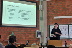 Lukas Baumann (Mann mit langen braunen Haaren, schwarzer Mütze und schwarzem T-Shirt) im Seminarraum neben einer projizierten Folie, auf der „Kontextfaktoren” zu lesen ist.