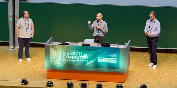 Im Hörsaal stehen drei Männer vor einem Schreibtisch, auf dem der Titel der Konferenz steht. Der Mann in der Mitte hält ein Mikrofon und spricht.