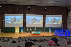 In einem großen Hörsaal mit drei Projektionsflächen stehen drei Männer auf dem Podium vor einem Tisch. Mehrere Anzeichen der Tagung sind zu erkennen, darunter beleuchtete Buchstaben mit der Aufschrift „MUC 26”.