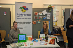 Ein Stand der Frühförderstelle im Kreis Unna zeigt Materialien zur Entwicklungshilfe für Kinder. Zwei Personen sitzen hinter dem Tisch, auf dem Broschüren, Arbeitsmaterialien und ein grünes Tablet zu sehen sind.