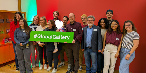 Eine Gruppe Personen schaut erfreut in Richtung Kamera und hält dabei ein grünes Schild mit dem Hashtag #GlobalGallery in weißer Schrift hoch.