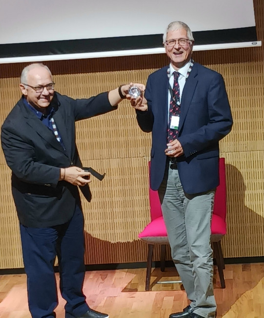 Auf dem Bild sieht man, wie der Award an Herrn Bühler vergeben wird. Er und ein weiterer Mann schauen in die Kamera und halten den Award in den Händen.