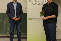 Zwei Personen stehen in einem Seminarraum vor einer Tafel und einem Roll-up-Banner. Auf dem Banner steht „70 Jahre rehawiss – Der Blick zurück nach vorne“. Eine Person hält eine kleine Topfpflanze in der Hand und trägt ein dunkles Hemd, eine blaue Hose und Turnschuhe. Die andere Person trägt einen Blazer, ein hellblaues Hemd, Jeans und schwarze Schuhe. Im Hintergrund ist ein Teil einer Präsentationsfolie mit dem Namen „Lena Hünefeld” und dem Datum „20.05.2025” zu sehen.