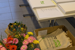 Ein Tisch mit mehreren weißen Mappen, die das grüne Logo der Technischen Universität Dortmund tragen. Die Mappen sind ordentlich in Reihen ausgelegt und scheinen für eine Veranstaltung vorbereitet zu sein. Daneben steht ein großer Eimer mit bunten Blumen in Gelb, Rosa, Orange und Rot sowie eine offene Kartonbox mit Stofftaschen, auf denen der Schriftzug „70 Jahre Reha-Wissenschaften“ zu lesen ist.