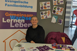 Eine Person sitzt hinter einem Tisch am Stand der Evangelischen Familienbildung Recklinghausen (Diakonie). Auf dem Tisch liegen Flyer, Broschüren und kleine Giveaways; daneben steht ein großes Banner mit Schriftzug „Gemeinsam wachsen Lernen Leben“.