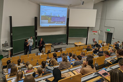 Blick über die Köpfe von Studierenden in einem Hörsaal nach vorne. Vorne stehen zwei Frauen und ein Mann un blicken in Richtung der Studierenden. Auf einer großen Leinwand ist eine Internetseite der Werkstatt über den Teichen zu sehen.
