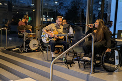 Eine kleine Musikgruppe spielt auf einer erhöhten Fläche. Eine Person sitzt am Schlagzeug, eine andere spielt Gitarre und eine weitere Person singt ins Mikrofon, während sie im Rollstuhl sitzt.