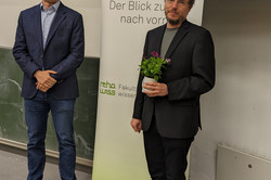 Zwei Personen stehen in einem Vorlesungssaal vor einer Tafel. Eine trägt einen dunklen Blazer mit Jeans, die andere einen schwarzen Anzug mit grauem Shirt und hält eine kleine Pflanze in den Händen. Hinter ihnen ist ein Roll-Up-Banner mit der Aufschrift „70 Jahre rehawiss – Der Blick zurück nach vorn“ zu sehen, ergänzt durch „Fakultät Rehabilitationswissenschaften“. Im Hintergrund ist eine Präsentationsfolie sichtbar, auf der unter anderem „Matthias R. Hastall“ und das Datum „20.05.2025“ stehen.
