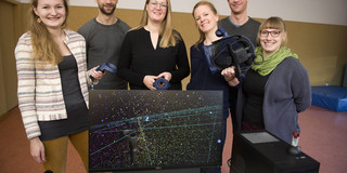 Mitarbeitende des Forschungsprojektes "Virtual Reality Moves" (VRM) stehen hinter einem Computer-Bildschirm, eine Person hält ein Virtual Reality Headset in der Hand 