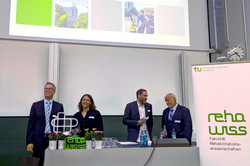 Das Bild zeigt den Hörsaal. Dort ist eine Präsentation auf der Leinwand zu sehen, die den Titel "Der Blick zurück nach vorne" trägt und verschiedene Bilder zeigt. Vor der Leinwand stehen mehrere Personen an einem Pult. Rechts im Bild steht ein Banner mit dem Logo der Fakultät für Rehabilitationswissenschaften.