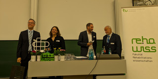 Das Bild zeigt den Hörsaal. Dort ist eine Präsentation auf der Leinwand zu sehen, die den Titel "Der Blick zurück nach vorne" trägt und verschiedene Bilder zeigt. Vor der Leinwand stehen mehrere Personen an einem Pult. Rechts im Bild steht ein Banner mit dem Logo der Fakultät für Rehabilitationswissenschaften.