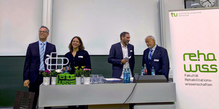 Das Bild zeigt den Hörsaal. Dort ist eine Präsentation auf der Leinwand zu sehen, die den Titel "Der Blick zurück nach vorne" trägt und verschiedene Bilder zeigt. Vor der Leinwand stehen mehrere Personen an einem Pult. Rechts im Bild steht ein Banner mit dem Logo der Fakultät für Rehabilitationswissenschaften.