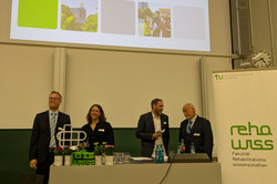 Das Bild zeigt den Hörsaal. Dort ist eine Präsentation auf der Leinwand zu sehen, die den Titel "Der Blick zurück nach vorne" trägt und verschiedene Bilder zeigt. Vor der Leinwand stehen mehrere Personen an einem Pult. Rechts im Bild steht ein Banner mit dem Logo der Fakultät für Rehabilitationswissenschaften.