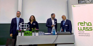 Das Bild zeigt den Hörsaal. Dort ist eine Präsentation auf der Leinwand zu sehen, die den Titel "Der Blick zurück nach vorne" trägt und verschiedene Bilder zeigt. Vor der Leinwand stehen mehrere Personen an einem Pult. Rechts im Bild steht ein Banner mit dem Logo der Fakultät für Rehabilitationswissenschaften.