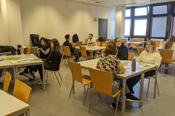 In einem Seminarraum sitzen mehrere Gruppen von Menschen an Tischen bei Gesprächen oder Diskussionen.