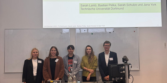 Drei Frauen und zwei Männer stehen hinter einem Hörsaalpult und schauen in Richtung Kamera.