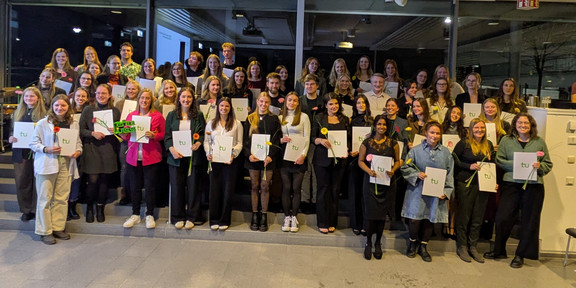 Eine große Gruppe von Personen steht in mehreren Reihen in einem hellen Raum der Technischen Universität Dortmund und machen ein Gruppenfoto. Viele halten Mappen mit dem grünen TU-Logo und einzelne Blumen in den Händen.