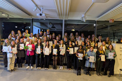 Eine große Gruppe von Personen steht in mehreren Reihen in einem hellen Raum der Technischen Universität Dortmund und machen ein Gruppenfoto. Viele halten Mappen mit dem grünen TU-Logo und einzelne Blumen in den Händen.