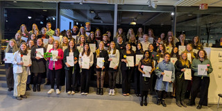 Eine große Gruppe von Personen steht in mehreren Reihen in einem hellen Raum der Technischen Universität Dortmund und machen ein Gruppenfoto. Viele halten Mappen mit dem grünen TU-Logo und einzelne Blumen in den Händen.