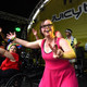 Man sieht eine Band auf einer Bühne. Eine junge Frau in pinkem Kleid zeigt mit ihren Händen ein "Peace"-Zeichen in die Kamera. Neben ihr sitzt eine Frau im Rollstuhl und hält ein Mikrofon in der Hand.