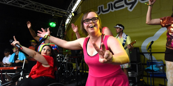Man sieht eine Band auf einer Bühne. Eine junge Frau in pinkem Kleid zeigt mit ihren Händen ein "Peace"-Zeichen in die Kamera. Neben ihr sitzt eine Frau im Rollstuhl und hält ein Mikrofon in der Hand.