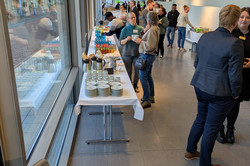 Ein langer Gang mit Fensterfront zeigt mehrere Tische voller Getränke, Tellerstapel und Snacks auf weißer Tischdecke. Menschen stehen zusammen, essen oder unterhalten sich.