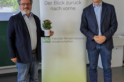 Zwei Männer stehen vor dem Roll-up der Veranstaltung „70 Jahre rehawiss – Der Blick zurück nach vorne“. Der Mann links trägt ein dunkelblaues Sakko, ein hellblaues Hemd, Jeans und braune Schuhe. Er hält eine weiße Tasse mit einer kleinen Pflanze. Der Mann rechts trägt ebenfalls ein dunkelblaues Sakko, hellblaues Hemd und Jeans und steht mit gefalteten Händen lächelnd daneben.