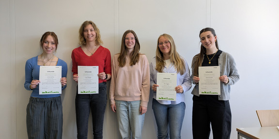 Fünf Frauen stehen nebeneinander von einer weißen Wand. Sie lächeln und halten Urkunden in die Kamera.