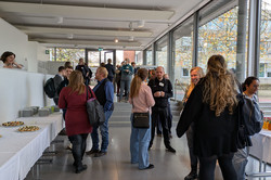 Menschen stehen und unterhalten sich im Foyer einer Veranstaltung. Links sind Tische mit weißen Tischdecken aufgebaut, darauf Tellerstapel und kleine Snacks.