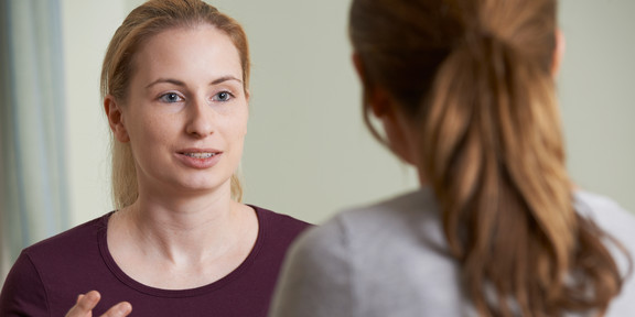 Zwei Frauen unterhalten sich. Eine braunhaarige Frau ist mit dem Hinterkopf zur Kamera gerichtet und eine blonde Frau ist mit dem Gesicht zur Kamera gerichtet, während sie der anderen Frau mit Hilfe von Handgestiken etwas erzählt.