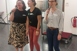Auf dem Bild sind drei Frauen zu sehen, die Mikrofone in den Händen halten. Sie singen Karaoke. Die Frau links trägt einen langen Rock mit einem Pflanzenmuster und ein schwarzes T-Shirt, während die Frau in der Mitte ein schwarzes T-Shirt und eine rote Hose trägt. Die dritte Frau rechts hat ein gestreiftes Hemd an und trägt Jeans.