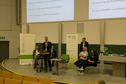 Das Bild zeigt den Hörsaal, in dem 6 Menschen vorne stehen und etwas präsentieren. Eine Person spricht in ein Mikrofon. Die anderen Personen sitzen oder stehen um die Person herum und hören zu. Im Hintergrund sind 2 große Leinwände zu sehen. Darauf werden die verschiedenen Jubiläen im Bereich der Sonderpädagogik und Rehabilitationswissenschaften angezeigt, die gefeiert werden. 