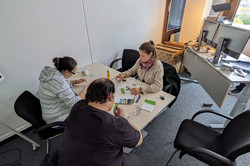 Blick von schräg oben auf einen Tisch an dem zwei Frauen und ein Mann Materialien an Materialien für einen Schaukasten arbeiten. Im Hintergrund steht ein Schreibtisch mit Computerbildschirm.
