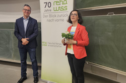 Zwei Personen stehen nebeneinander in einem Seminarraum vor einer Tafel. Eine Person trägt einen roten Blazer und hält einen kleinen Blumenstrauß in den Händen. Hinter ihnen ist ein Roll-Up-Banner mit der Aufschrift „70 Jahre rehawiss – Der Blick zurück nach vorn“ zu sehen, darunter die Ergänzung „Fakultät Rehabilitationswissenschaften“. 