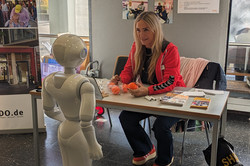 Eine Person sitzt an einem Informationstisch des WBZ Zehnthof („Junge Pflege“) gegenüber eines humanoiden Roboters, der scheinbar interagiert oder kommuniziert