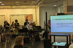 Die Musikgruppe der musikalischen Mittagspause mit etwa zehn Personen tritt im Foyer der EF50 auf. Die Mitglieder spielen verschiedene Instrumente. Einige singen. Rechts im Bild ist eine große Leinwand zu sehen mit dem Text: „Die Musikalische Mittagspause begrüßt Sie herzlich und freut sich auf Ihre gesangsstarke Unterstützung“. Im Vordergrund sitzen und stehen einige Zuschauer*innen.