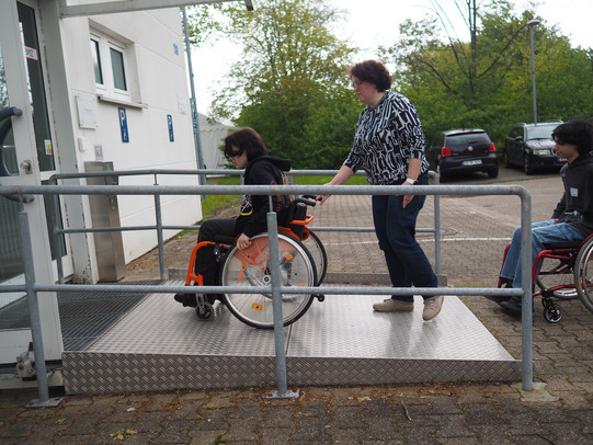 Ein Junge sitzt auf einem Rollstuhl und fährt eine Rampe hoch. Hinter ihm steht eine Frau und hält ihre Hand an den Rollstuhl.