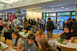 Das Bild zeigt das Foyer des EF50, in dem mehrere Personen an Tischen sitzen und sich unterhalten. Die Tische sind mit weißen Tischdecken und teilweise mit Dekoration wie Blumen und Getränken versehen. Einige Personen stehen im Hintergrund am Buffet, während die meisten sitzen, essen und sich unterhalten.