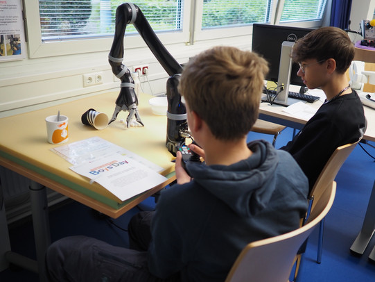 Zwei Jungen sitzen an einem Tisch, ein Roboterarm mit 3 Fingern greift auf den Tisch.