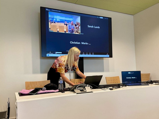 Eine blonde Frau schaut im Computer und steht in ein Podium. Im Hintergrund läuft ein Zoom-Meeting.
