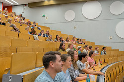 Auf dem Bild sind die Sitzreihen des Hörsaals zu sehen, in den viele Menschen sitzen. Einige haben Getränke auf den Tischflächen vor sich stehen. Sie scheinen aufmerksam zuzuhören.