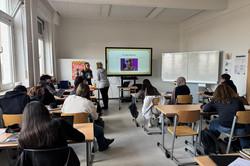 Das Bild zeigt einen Klassenraum, in dem eine Lehrveranstaltung stattfindet. Im Vordergrund sitzen mehrere Schüler*innen an Tischen, einige von ihnen arbeiten an Laptops oder Notizen. An der Wand ist ein großer Bildschirm installiert, auf dem das Thema „KI oder Mensch“ präsentiert wird. Eine Frau steht vor der Klasse und präsentiert den Inhalt. 
