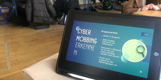 Das Bild zeigt einen Unterrichtsraum, in dem Schüler*innen an Tischen sitzen und an einer Gruppenarbeit teilnehmen. Im Vordergrund ist ein Tablet zu sehen, das die Anweisungen für die Gruppenarbeit zum Thema „Cybermobbing erkennen“ anzeigt. Die Aufgaben beinhalten die Bildung von vier Gruppen, wobei jede Gruppe ein Fallbeispiel erhält, das sie diskutieren soll.