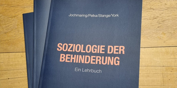 Drei Bücher liegen auf einem Tisch. Titel des oberen Buches ist "Soziologie der Behinderung: Ein Lehrbuch" in orangener Schrift auf schwarzem Grund. Darüber die NAmen der Autor*innen Jochmaring, Pelka, Stange und York.