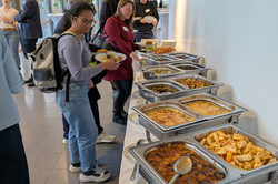 Mehrere Personen bedienen sich an einem warmen Buffet mit verschiedenen Speisen in Edelstahlbehältern. Andere Gäste unterhalten sich im Hintergrund bei Getränken.