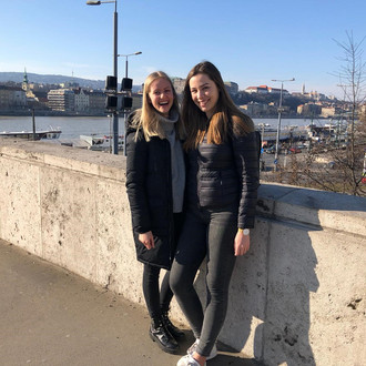 Zwei Studentinnen mit Fakultätslogo auf einer Flussbrücke in Budapest