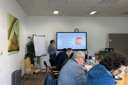 Das Bild zeigt einen Schulungsraum, in dem eine Präsentation stattfindet. An einem langen Tisch sitzen mehrere Teilnehmer*innen, die auf den Bildschirm vor ihnen schauen. Der Bildschirm zeigt das Thema „Fragebogen 1.1“ und ein Stoppschild-Symbol. Eine Person steht vorne im Raum und erläutert den Inhalt der Präsentation. Neben dem Tisch befinden sich Flipchart-Papier und einige Pflanzen. 